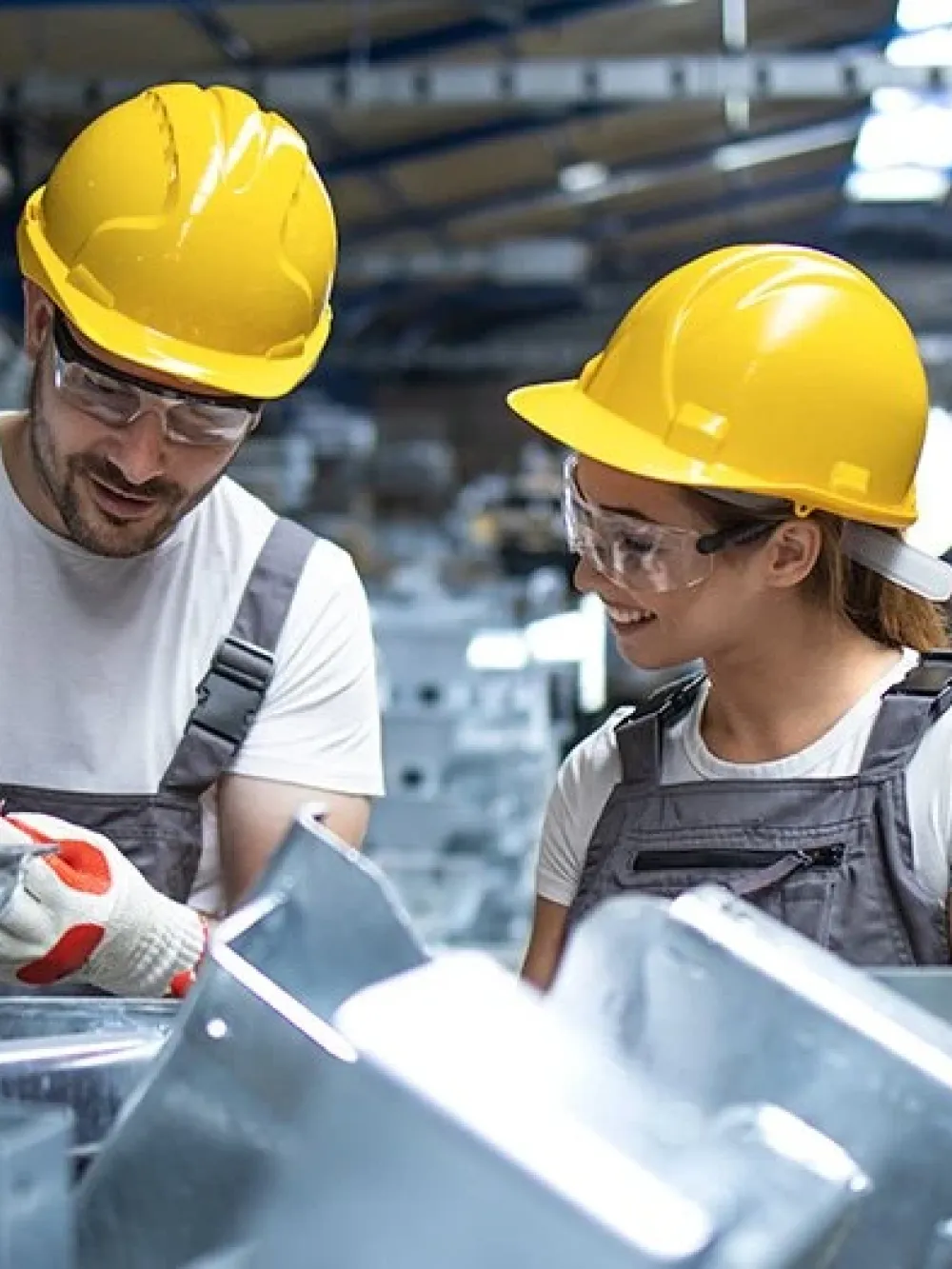 Deux personnes portant des casques de sécurité jaunes travaillent avec des pièces métalliques dans un atelier industriel.